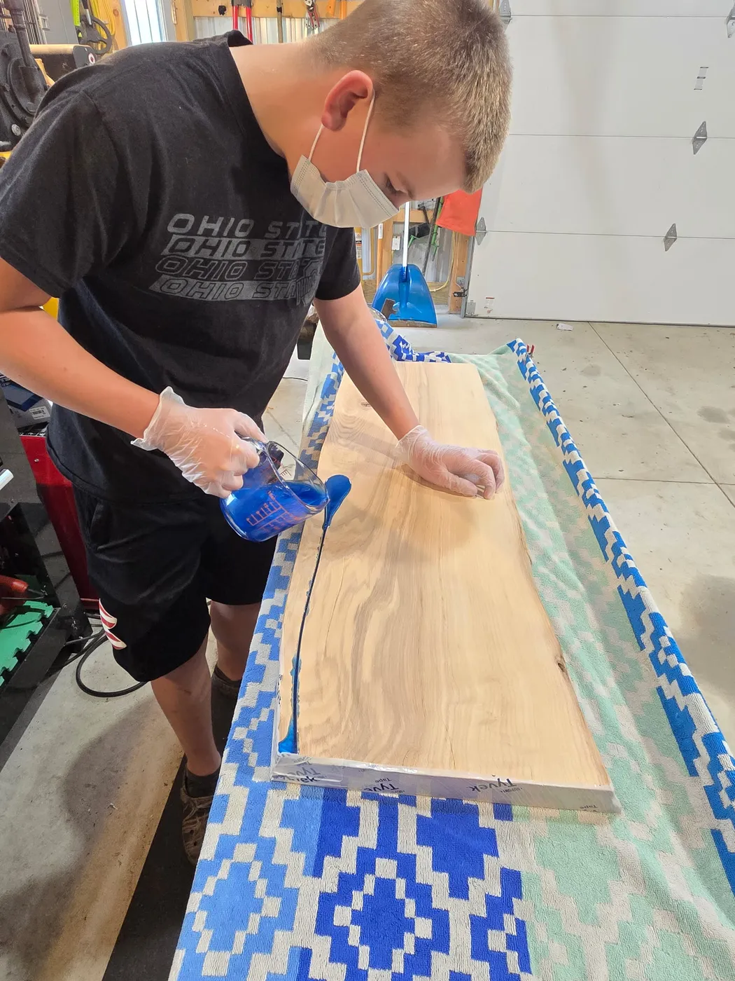 Brody pouring pigmented epoxy along the edge of a live edge slab in his shop